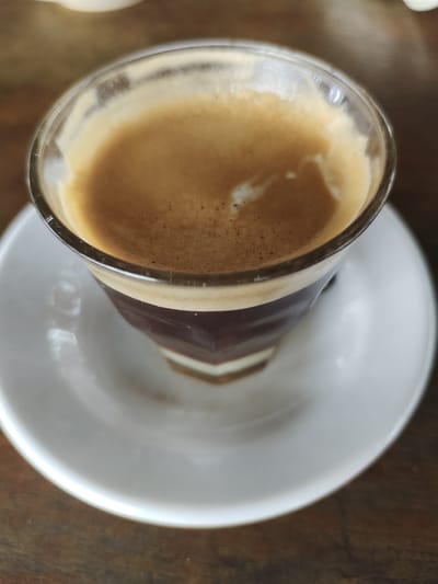 A close-up, high-angle shot shows a clear glass of espresso with a thick layer of crema, resting on a white saucer. The coffee is dark brown with lighter brown foam on top, displaying a swirling pattern. The glass has a faceted design, and the saucer has a subtle curve. The background is a blurred wooden surface, suggesting an indoor setting. The image was taken in Keudematangkuli, Indonesia. There are no people visible in the scene.