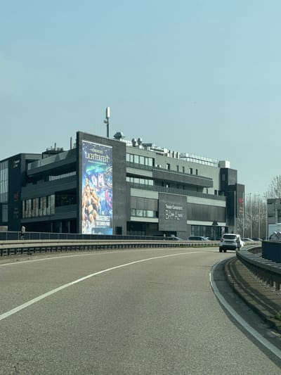The image displays a modern, multi-story commercial building situated adjacent to a multi-lane asphalt road, in the vicinity of Altenheim, Germany. The building features a dark grey or black facade with numerous windows and recessed sections, suggesting balconies or additional architectural elements. A large vertical banner on the left side of the building advertises "Nordisches Lichterfest" and "Wasserwelt des Europa-Park," depicting individuals in swimwear alongside illuminated scenery. To the right, text signage on the building reads "Theater Eurodistrict BAden ALsace." The rooftop contains various mechanical installations, including ventilation units and communication antennas. In the foreground, a two-lane road with a solid white center line curves to the right, bordered by a metal guardrail. A white passenger vehicle is observed traveling in the right lane, moving away from the viewer. Several other vehicles are visible parked or in the distance behind the guardrail. The sky is clear and light blue, with some bare trees visible in the far background to the right.