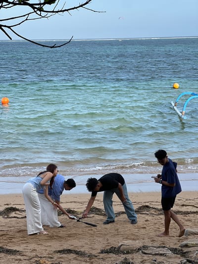 Four individuals are gathered on a sandy beach near the ocean in Denpasar, Indonesia. A woman wearing a light blue top and white pants is bending over, looking at something on the ground. A man in a blue shirt and dark pants is also bending down, holding a phone and a selfie stick. Another man in a black shirt and light blue pants is bent over as well, seemingly interacting with something on the sand. A fourth individual, a young man in a dark blue shirt and black shorts, stands to the right, holding a phone and looking down at it. The ocean stretches out behind them, with waves gently lapping the shore. In the water, there are two buoys, one orange and one blue, and a kite is visible in the sky. The beach is covered in sand and scattered with seaweed and rocks.