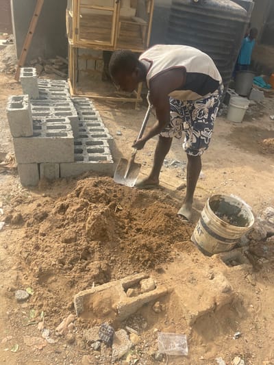 A man mixes soil with a shovel, next to a pile of gray cinder blocks. He wears a white shirt, patterned shorts, and sandals. A bucket sits nearby, its contents possibly related to the soil mixing. In the background, a wooden structure stands beside what appears to be a building with other people nearby. The scene seems to be a construction site or a home improvement project.
