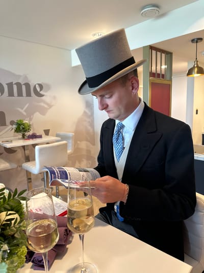 An indoor scene in Epsom and Ewell, United Kingdom, depicts a Caucasian man, appearing to be in his 20s or 30s, seated at a white table, engrossed in reading. He is dressed in formal morning wear, including a light grey top hat with a black band, a white collared shirt, a light blue patterned tie, a light-colored waistcoat, and a dark (likely black) tailcoat. A small gold hoop earring is visible in his left ear, and a thin red string bracelet along with a gold chain bracelet adorn his left wrist.

On the white table in the foreground, two champagne flutes filled with a sparkling golden liquid are positioned. A small floral arrangement featuring green foliage and white flowers sits to the left of the flutes, next to a stack of purple folded napkins. The man holds an open program or newspaper with the words "RACE SUMMARY" clearly visible, suggesting it is a race card or betting guide.

The background shows a brightly lit room with white walls and ceiling. A large grey graphic on the wall to the left partially displays the text "me" and a faint silhouette of a person wearing a hat. Further back, a tall, narrow white table with two white cushioned bar stools is visible. A smaller floral arrangement and purple folded fabric are on this table. Architectural elements include a light green framed partition with a dark red panel. On the white ceiling, a circular white smoke detector is visible, along with a recessed light fixture. A brass-colored pendant lamp with a warm glow hangs from the ceiling on the right side. The overall environment suggests a hospitality suite or private lounge at a racecourse, consistent with Epsom Downs Racecourse.