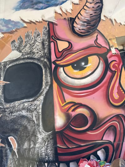 This is a close-up of a partially damaged piece of street art, presumably a mural on a wall. The top half of the mural features a stylized eye with a yellow iris, black pupil, and red and pink surrounding shapes and lines. The bottom half is a black and white drawing with a large black shape resembling a nose. The surrounding area of the "nose" is filled with smaller, detailed drawings. The mural is torn in places, revealing a light brown surface underneath. In the upper right corner, there is a glimpse of what appears to be a white object with some colorful fabrics draped on it. The scene is located in Salta, Argentina.