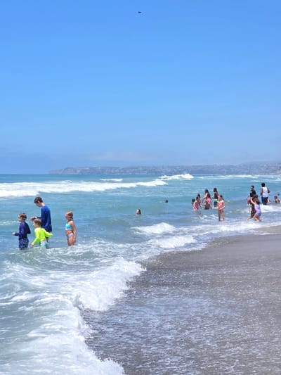 The image depicts a sunny day at a beach in San Clemente, California. Several people, predominantly children, are in the ocean wading and playing in the waves. In the foreground, a group of children are standing in the shallow water. To the left, a young boy in a blue rash guard and patterned swim trunks stands with a person in a blue shirt. Next to him is another boy in a bright yellow rash guard and green swim trunks. A girl in a blue bikini top and patterned bottoms, wearing pink goggles, stands to the right of the boys. In the mid-ground, more people are scattered in the water, some swimming and others standing. Further in the background, the coastline of San Clemente is visible with houses and hills. The ocean is a clear blue-green with white-capped waves rolling towards the shore. The sky is a bright, cloudless blue. A small dark object is visible in the upper center of the sky.
