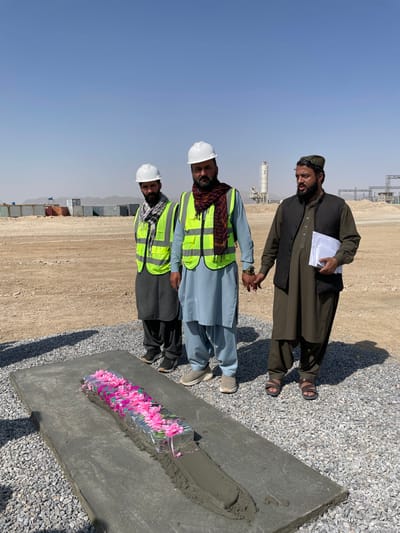 The image depicts three adult males standing on a gravel surface in Pir Dust, Afghanistan, under a clear blue sky at what appears to be a construction or industrial site. In the foreground, a rectangular concrete slab lies horizontally on the ground. A central trough in the slab contains fresh, wet cement, into which a long, narrow object wrapped in reflective silver material and adorned with numerous bright pink artificial flowers or petals has been placed.
The three men are positioned behind and slightly to the right of this slab. The two men on the left wear white hard hats and neon yellow safety vests over traditional attire; the middle man also wears a multi-colored scarf. The man on the far right wears a dark waistcoat over olive-green traditional clothing and a dark cap, holding a stack of white papers in his left hand. The man in the center and the man on the right are holding hands.
In the background, a flat, dusty landscape extends to a low horizon of hills. Various industrial elements are visible, including multiple shipping containers, tall silos or towers resembling a concrete batching plant, and utility poles or transmission structures. The ground directly beneath the men and the slab is covered in grey gravel, transitioning to dirt and sand further into the background. Shadows indicate direct sunlight.
