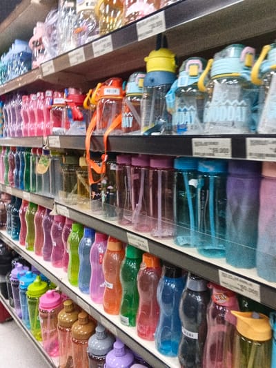 Shelves stocked with an array of colorful water bottles are displayed in a retail setting. The bottles vary in shape and size, featuring hues of pink, purple, green, blue, orange, and clear. Some have straws and carrying straps. Price tags are visible on the shelves, suggesting these are products for sale. The lighting in the store illuminates the diverse selection of bottles.