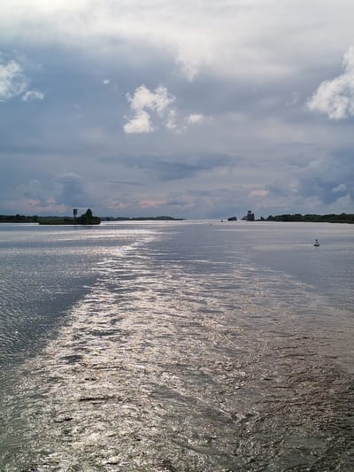 The image shows a wide, calm body of water under a cloudy sky. The water reflects the light, creating a shimmering effect. In the distance, there is a treeline and a few structures, including what appears to be a lighthouse or tower. There are also a few buoys in the water. The overall scene suggests a tranquil, somewhat overcast day on a river or bay. There is no indication of the city of Kargulino, Russia in this image.