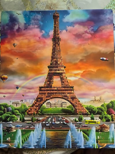This image depicts a surreal, dreamlike cityscape with a colossal Eiffel Tower constructed from an assortment of round, jewel-like objects in various shades of brown and pink. The tower stands prominently in the foreground, with an arched opening at its base revealing a quaint, European-style building. Surrounding the tower is a lush green landscape with manicured trees and a vibrant flowerbed. In the distance, several architectural landmarks are visible, including what appears to be the Arc de Triomphe and a domed building reminiscent of Sacré-Cœur Basilica, suggesting a Parisian setting. The sky is a dramatic gradient of orange, pink, and blue, filled with fluffy clouds, hot air balloons, and a dirigible. A rainbow arcs gracefully across the sky. In the foreground, a series of fountains spray water, and a reflective pool mirrors the scene. Miniature figures and vehicles are scattered throughout the landscape, adding a sense of scale and activity. The image does not contain any elements related to Serebriansk, Kazakhstan.