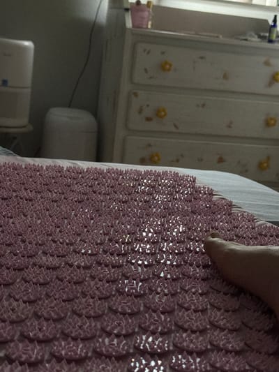 The image shows a human foot resting on a pink spiky mat. The mat has a repeating pattern of small, raised, spiky elements. In the background, there is a light-colored cabinet or closet with floral patterns. Below the cabinet, a portion of a white cylindrical object and a small, rectangular object are visible.
While a location within Morton Grove, United States of America, cannot be determined from the image alone, the scene appears to be indoors, likely in a bedroom or similar personal space.