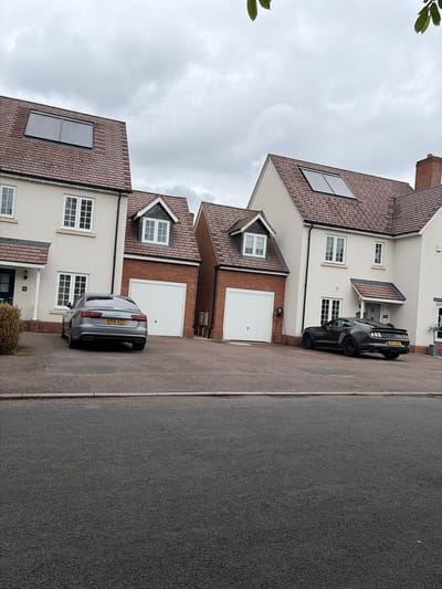 This image depicts a residential street scene in Leicester, United Kingdom, under an overcast sky. Two distinct houses, numbered 28a and 28b, dominate the midground, each with an attached garage and individual driveways.
The house on the left (28a) features a light-colored rendered facade above a red-brick lower section, a dark blue front door with a small canopy, and white-framed windows, some with internal white louvered shutters. Its pitched roof is covered with red-brown tiles and includes a rectangular, dark gray solar panel array. An integrated garage with a white roller door is visible on the right side of the house. A silver-grey Audi A6 sedan, with license plate "KP16 XZT", is parked facing away from the camera in its driveway.
The house on the right (28b) mirrors the architectural style of 28a, featuring a light-colored rendered facade, red-brick elements, and a grey front door with a canopy. Its tiled roof also has a similar dark gray solar panel array and a red-brick chimney on the right side. An integrated garage with a white roller door is present, and an electric vehicle charging point is mounted on the wall to the left of the garage door. A dark grey Ford Mustang, identifiable by its rear spoiler and quad exhaust pipes, with license plate "MDD3 HAT", is parked facing away from the camera in its driveway, positioned close to the house and partially obscuring the charging point.
Both houses display dormer windows above their respective garages. The driveways are paved with dark material. In the foreground, a dark asphalt road is separated from the driveways by a light grey curb. No individuals are visible in the scene. The sky is uniformly covered with grey clouds. Minimal green foliage is visible at the very top edge of the frame.