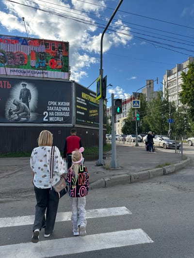The image shows a street scene in Rubtsovo, Russia, on a sunny day with scattered clouds. The perspective is from behind a group of people walking on a crosswalk, looking towards the street and buildings.
In the foreground, three people are visible from the waist down, walking on a zebra crossing. The person closest to the camera is wearing a white shirt with a polka dot pattern and dark pants. The middle person is wearing dark pants and a light-colored shirt. The person furthest away is wearing a black jacket and possibly dark pants. Two of them have bags.
The scene depicts a street with a sidewalk, a paved road, and buildings in the background. Traffic lights are visible, with one showing a green pedestrian signal. A large billboard with colorful advertising is on the left side of the image. Another billboard displays text in Russian. The general atmosphere appears to be one of everyday urban life. The time of day is likely daytime, given the bright sunlight and clear sky. There are no obvious indications of any specific event or strong emotions. The text on the billboards is in Russian, with one legible phrase appearing to be "Срок жизни закладчика на свободе 2 месяца," which translates to "The jail time for a drug dealer is 2 months."