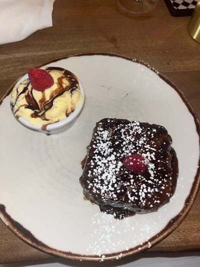 A close-up, overhead shot displays two dessert items on a round, off-white ceramic plate with a rustic brown rim, resting on a light brown wooden table. On the left, a small white ramekin contains light yellow ice cream or custard, generously drizzled with dark chocolate sauce and topped with a single red raspberry. To the right, a rectangular dark chocolate dessert, possibly a brownie or cake, is covered in a glossy dark chocolate glaze, dusted with white powdered sugar, and garnished with a single red raspberry. A blurred wine glass is partially visible in the upper right background, and a crumpled white napkin is present in the upper left. No individuals are depicted. The specific location within Stevenage, United Kingdom, cannot be determined from the visual information provided.