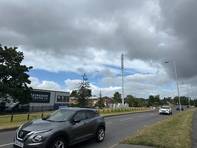 This media file captures a street scene in Reading, United Kingdom, during daytime under a sky featuring a mix of heavy grey clouds and patches of bright blue. The setting appears to be a commercial or light industrial area alongside a multi-lane road.
In the foreground, a dark grey Nissan Juke SUV, bearing the UK license plate "M100 BEG", is driving on the left side of the road, moving towards the viewer. Behind it, on the left, a large, dark green tree partially obscures a commercial building. This building has a prominent sign that reads "KINGDOM COFFEE CO" in large white letters. A black van with the "KINGDOM COFFEE CO" logo is parked behind a fence in front of the building. Another sign nearby indicates "Open to Trade & Public".
Further down the road, on the right side of the image, a tall, slender telecommunications mast stands against the sky. The road itself features white lane markings and is lined with streetlights. Several other vehicles are visible in the distance, including a white SUV. The surrounding landscape includes grassy verges, smaller trees, and more buildings further in the background. The weather conditions suggest a day of variable cloudiness, with sunlight breaking through in some areas.