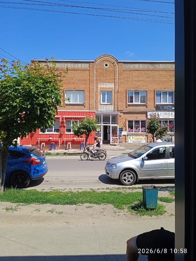 On a sunny day in the city of Lebedyn, Ukraine, a street scene unfolds. A blue car is parked on the left side of the road, facing away from the camera. A silver sedan is parked on the right side of the road, facing the camera. A person is riding a moped down the street between the two cars, moving from left to right. The road is paved, with a grassy verge on the left and a sidewalk on the right. A brick building with a red facade is visible behind the blue car and the moped rider. The building has several windows and doors. A tree with green leaves is in front of the building. The sky is clear and blue. A trash can is located on the sidewalk. The date and time 2026/6/8 10:58 are visible in the lower right corner of the image.