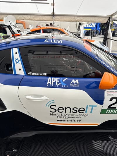 A blue and white Porsche race car with the number "2" on its side is parked under a tent in Stavelot, Belgium, near the coordinates 50.441 latitude and 5.969 longitude. The car has several sponsor logos on its side, including "APEX MARKETING", "MELAMED MOTORSPORT", and "Sense IT". The car has a Star of David sticker on its side. There are other race cars in the background. The car is parked on black and white checkered asphalt. The tent is white and has a red and white "RECLIN" sign on the side. There is a white truck in the background. The car is under a large white tent.