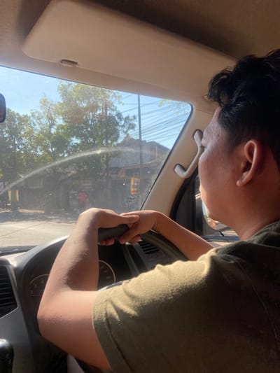 A man with dark, short hair is seen from the side, driving a vehicle. He is wearing a dark green t-shirt and has his hands on the steering wheel, which has a speedometer visible on the dashboard. The sun is shining brightly, and a spray of water is visible across the windshield. Outside the car, trees and buildings can be seen lining a street. The interior of the car shows a beige sun visor and a grab handle. The scene is likely in Jabon, Indonesia, given the presence of trees, buildings, and general streetscape characteristic of the region.