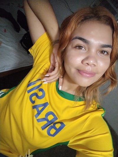 A young woman with reddish-brown hair and light brown eyes poses for a selfie. She is wearing a yellow t-shirt with blue lettering that appears to spell out "BRASIL." The t-shirt has green trim around the neckline and sleeves. Her right arm is bent behind her head, and her left hand is resting near her face. She is wearing hoop earrings and has white nail polish on her fingernails. The background is out of focus and appears to be an indoor setting with some dark objects, possibly clothing, on a surface. The lighting suggests it is daytime. There is no identifiable information within the image to confirm or deny a specific location in Punta de Piedras, Venezuela.