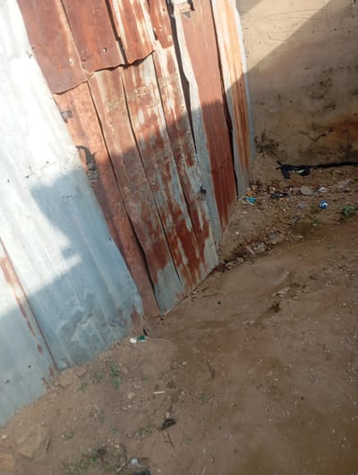 A vertical, close-up, eye-level, outdoor shot shows a weathered, corrugated metal wall constructed from sheets of rusty brown and silver-grey metal. The wall is situated in an urban environment in Gwagwa, Nigeria, with a dirt ground in the foreground and a rough, textured wall to the right. A pile of dark-colored plastic bags is visible in the mid-ground to the right of the metal wall, amidst scattered debris. The ground is uneven, with patches of dry soil and small stones. The overall scene depicts a neglected or informal area.