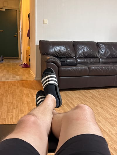 The image shows a person's legs and feet, wearing black Adidas slides with white stripes and black socks. The person is sitting on a floor with a wood-grain pattern. To the left, a dark brown leather sofa is visible, with a white wall behind it. A light switch is on the wall. Below the sofa, a doorway leads to another room with a dark green door. Clothing, including a purple garment and a black item, is piled near the doorway. The scene appears to be indoors, likely in a home. The lighting suggests it could be daytime or evening, with artificial light visible from the doorway. There is no visible text in the image.