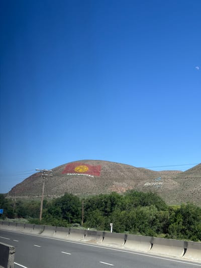 The image captures a mountainous landscape in Kyrgyzstan, as indicated by the visible text "КЫРГЫЗСТАН" and a large rendition of the Kyrgyz flag painted on the mountainside. The flag, featuring a yellow sun with a stylized crown in the center, is prominently displayed on the textured, arid slopes of the mountain. To the right of the mountain, a paved road runs alongside, bordered by a concrete barrier and lined with vegetation. The scene is bathed in bright sunlight, suggesting a clear day, likely mid-morning or afternoon given the strong shadows and the high position of the moon, which is faintly visible in the bright blue sky. Power lines and poles are scattered across the landscape, indicating infrastructure development in the area. The overall impression is one of a grand, natural setting with human elements like the road, flag, and text art integrated into it, showcasing national pride or a landmark. The lack of people suggests a focus on the landscape and its features.