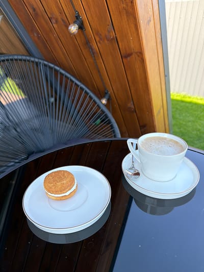 A white coffee cup with a thin gold rim sits on a matching saucer with a gold rim, accompanied by a silver spoon. The cup contains a frothy, light brown beverage. To the left, on a separate white plate with a gold rim, rests a single sandwich cookie with a beige cookie on top and bottom, and a white cream filling in between. Both plates and the cup are positioned on a dark, reflective surface, likely a table. In the background, a black wire chair with a fan-like back is visible. Behind the chair, a wooden structure with horizontal planks is present, and string lights with exposed bulbs are attached to it. A section of a white corrugated fence is to the right, with a green lawn extending beyond it. The scene suggests an outdoor setting, possibly a patio or garden area.
