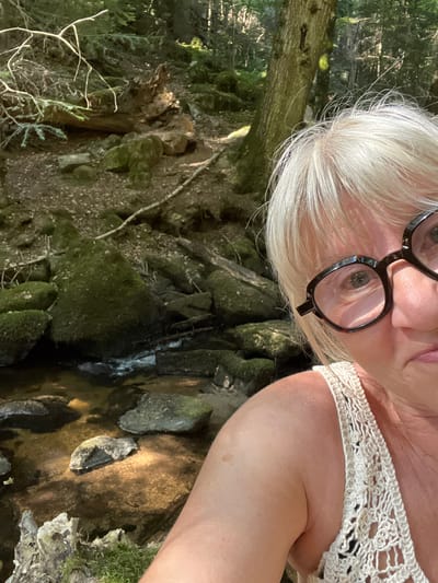 The image shows a woman with blonde hair and glasses, taking a selfie in a forest setting. She is wearing a white crocheted top. In the background, there is a stream or brook flowing over moss-covered rocks, surrounded by trees and foliage. The scene appears to be a natural, wooded area, possibly a hiking trail. The location is specified as Gumières, France.