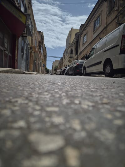 The image captures a street scene in Pietà, Malta, looking down from a low angle towards the sky. The foreground is dominated by the textured asphalt of the road, with individual pebbles and aggregate clearly visible due to the close perspective. Flanking the road are multi-story buildings with balconies and windows characteristic of Maltese architecture. On the right side of the street, several cars are parked, including a white van in the foreground. The buildings exhibit signs of age and wear, with some facades appearing weathered and discolored. The sky above is partly cloudy, with distinct white clouds against a bright blue sky. The scene suggests a typical urban environment in Malta, likely during daylight hours.