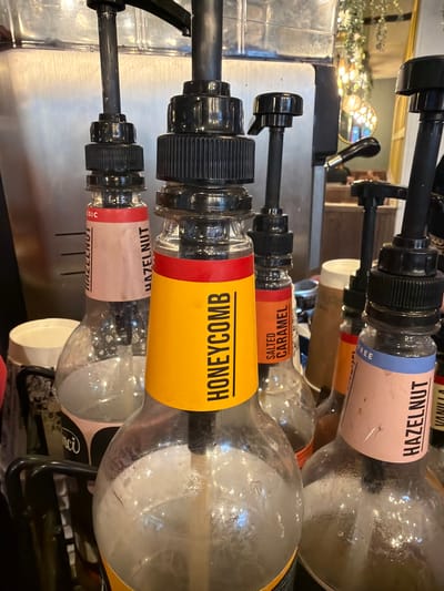 The image depicts a syrup dispensing station, likely within a cafe or coffee shop in Mole Valley, United Kingdom. Visible are four glass syrup bottles, each equipped with a dispensing pump. The syrup flavors are labeled as HAZELNUT, SALTED CARAMEL, and HONEYCOMB. The bottle labeled HONEYCOMB is the most prominent, with its yellow label and red accent. The other bottles are less focused and partially obscured. The dispensing pumps are black, and the bottles are supported by a metal frame. A stainless steel surface is visible, likely part of the counter.