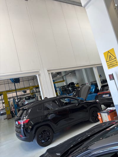 The image displays the interior of a car service center or repair shop, brightly lit with artificial lighting illuminating light-colored walls and a polished floor. In the foreground, a black SUV, resembling a Jeep Grand Cherokee, is prominently featured, with its rear section and a black multi-spoke wheel clearly visible. Another dark-colored vehicle is partially seen on the right edge.

The scene depicts car maintenance activities. In the mid-ground, a blue car has its hood open, positioned beneath a car lift, suggesting engine work. Further back, a silver compact car, possibly a Fiat, is elevated on a yellow hydraulic lift. Various pieces of workshop equipment, including other car lifts and mechanical tools, are scattered throughout the spacious area.

Notable details include a yellow triangular warning sign affixed to an overhead beam, displaying a forklift icon and the Italian text "ATTENZIONE CARRELLI IN MOVIMENTO," which translates to "Caution: Forklifts in motion." Below this, a red fire extinguisher is visible, labeled "ESTINTORE." The license plates on the vehicles, "HD-052ZB" on the black SUV and "EB 794GH" on the silver car, are in the Italian format, confirming the location context of Ancona, Italy. The time of day is not discernible due to the indoor setting, but the bright lighting suggests operational hours. No people are visible, and the overall atmosphere is functional and industrial.