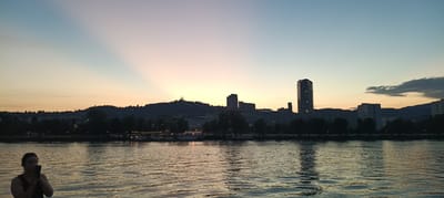The image captures a serene sunset over the Danube River in Linz, Austria, as viewed from the water. The sky transitions from a pale blue at the top to soft hues of pink and orange near the horizon, with prominent crepuscular rays emanating from behind a distant hill. Silhouetted against the sky is the cityscape of Linz, featuring a prominent hill topped with two church-like structures. Several modern buildings, including a distinctive tall, ribbed skyscraper, are visible along the riverbank. The river itself reflects the twilight sky and the city lights, creating a shimmering effect. In the foreground, a woman stands on a vessel, holding a smartphone and appearing to take a photograph or video of the scenery. The scene is peaceful, with the water providing a calm foreground to the impressive urban and natural panorama.