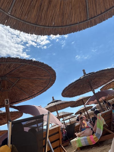 The image depicts a crowded beach scene under a bright blue sky with scattered clouds. In the foreground, the underside of a large thatched umbrella is visible. Several other similar umbrellas are scattered across the sandy beach, providing shade. People are seated on lounge chairs and are partially visible under the umbrellas. In the midground, a woman in a swimsuit is reclining on a striped towel-covered lounge chair, looking at her phone. Other beachgoers are also visible, some sitting, some standing, with a child playing nearby. A numbered sign "3" is attached to a wooden post supporting one of the umbrellas. The overall impression is a typical summer day at a busy beach resort. The location is identified as Nea Iraklitsa, Greece.