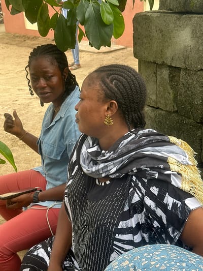 In Ganyingbo, Nigeria, two women are seated outdoors under the shade of tree foliage. The woman on the left, with braided hair and prominent side curls, wears a light blue denim shirt and coral-colored trousers. She holds a dark rectangular object, possibly a phone or tablet, in her left hand, and a thin, light-colored wire is visible connecting her to her lower body. She gestures with her right hand, palm facing outward, fingers slightly curled. The woman on the right, also with braided hair, is adorned with a gold leaf-shaped earring in her left ear and wears a black and white patterned top with a black and white scarf featuring gold fringing around her neck and shoulders. Her posture suggests she is looking to her right. Behind them, a rough stone wall is visible. The ground appears to be sandy soil. A person in the background, partially obscured by leaves, is wearing a blue shirt and light-colored trousers.