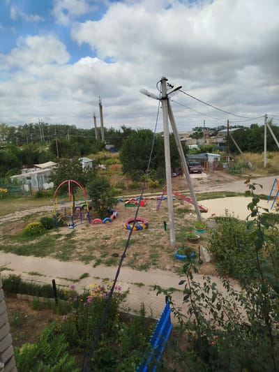 The image depicts a view of a residential area in the village of Kuzmichi, Russia, on a cloudy day. In the foreground, a section of a brick wall and dense green foliage are visible. A blue fence can be seen to the right. The middle ground features a playground with a colorful metal structure, a slide, and a sandbox. Tires have been repurposed as decorative elements and borders for the playground. A dirt path winds through the scene, and several power poles with overhead wires are prominent. In the background, trees, greenhouses, and a parked car are visible, along with other structures and utility poles, suggesting a semi-rural or suburban setting. The sky is filled with a mixture of white and gray clouds.