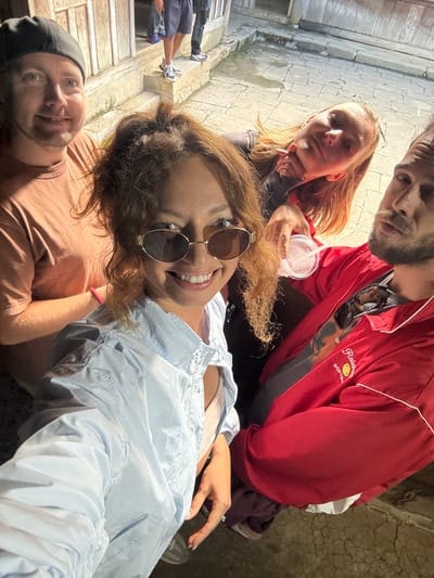 Four people are captured in a selfie taken outdoors in Sà Phìn Commune, Vietnam. The focal point is a woman in the center, wearing round sunglasses and a light blue jacket. To her right is a man in a red jacket, holding a plastic cup with a straw. To her left is a man wearing a black cap. Another woman is visible in the background, positioned behind the woman in sunglasses. Part of a stone structure, potentially a staircase or wall, is visible on the left. The ground is a combination of stone and dirt.