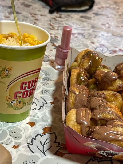 A top-down, rotated view depicts a cup of corn, a makeup item, and pastries in a cardboard container. The "CUP CORN" cup, featuring cartoon corn images, contains corn kernels and is being eaten with a utensil. A lip gloss product rests on the table. The pastries are topped with chocolate and a pistachio-colored glaze. All objects are placed on a table with a patterned surface. This image appears to be taken in Galiche, Bulgaria, based on visual features such as the product design of the "CUP CORN" cup.