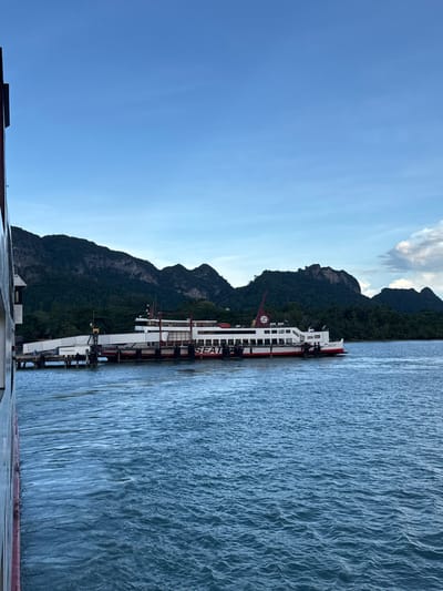 A ferry, identified by the text "SEAT" prominently displayed on its side, is docked at a pier under a cloudy sky. The ferry is white with red trim and has multiple decks. The pier is a long structure extending into the water, with various supports and railings. In the background, lush green mountains rise above a dense treeline, characteristic of the tropical landscape of Tambon Don Sak, Thailand. The water in the foreground is a deep blue, with gentle ripples. The left edge of the frame shows the side of another vessel, also with red trim. The scene suggests a travel hub or port in a scenic coastal region.