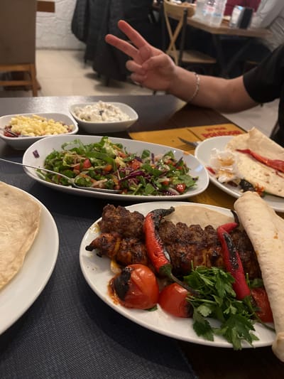 The image depicts an indoor restaurant setting, specifically identified as Pidom Burger & Köfte in Manavkuyu Mahallesi, Bayraklı, İzmir, Turkey. A dark-colored table, partially covered by a dark blue placemat, displays multiple dishes of food.
In the foreground, a white oval plate holds several pieces of grilled meat, possibly kebab or kofta, accompanied by two grilled red peppers, two grilled red tomatoes, and a sprig of fresh parsley. A piece of flatbread (lavash) is draped over a portion of this food. To its left, another white plate holds a piece of similar flatbread.
In the midground, a large white oval bowl contains a mixed green salad, including lettuce, red cabbage, and diced tomatoes, with a serving spoon. Behind it, two small white rectangular dishes are present: one filled with small white diced cubes, possibly cheese, and the other with a creamy, white dip containing darker elements, likely olives. A partially consumed dish with torn flatbread and red residue is visible to the right.
A person's right arm and hand are visible in the upper midground, extending from the right. The hand is displaying a "V" sign with the index and middle fingers extended. A silver-colored bracelet is visible on the person's wrist. The background features blurred elements of the restaurant interior, including wooden chairs, another table with water bottles, and indistinct patrons. A yellow placemat with the partial text "PİDOM" is visible under some dishes.