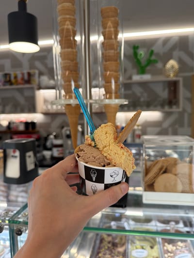A person is holding an ice cream cup in their hand, likely inside a gelato shop. The ice cream cup is decorated with black-and-white designs of cones and other ice cream treats. The cup contains two scoops of ice cream: a light brown scoop and a darker brown scoop. A small piece of wafer is stuck in the light brown scoop, and a blue plastic spoon is also inserted. Behind the ice cream, there's a container of waffle cones. The shop's interior includes a gelato display case with various flavors visible through the glass. The general location is Rosolini, Italy, indicated by the image's characteristics.