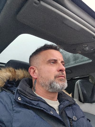 An adult male subject is depicted seated inside a vehicle in Itzig, Luxembourg. He is positioned in the foreground, facing right with a neutral expression. The male has short, dark hair with faded sides, a prominent grey beard, and dark eyes. Two small black hoop piercings are visible in his left ear, and a dark tattoo extends downwards on the left side of his neck. He wears a dark blue winter jacket with a brown fur-lined hood, over a light cream-colored long-sleeve top. A black seatbelt is visible across his chest. The vehicle's grey headliner is visible above, with a sunroof showing an overcast sky. The headrest of an adjacent seat is visible to the right of the subject.
