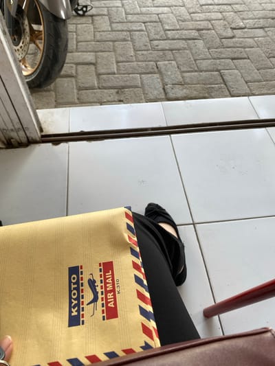 The image presents a low-angle view from the perspective of a person who appears to be seated or crouching, showing their legs and the immediate surroundings. In the foreground, a pair of legs dressed in black trousers or fabric is visible, with a hand holding a brown air mail envelope against the thigh. The envelope features distinctive red, white, and blue striped borders. Visible text on the envelope, oriented upside down from this perspective, reads "AIR MAIL" and "K.310", with "KYOTO" printed below an airplane graphic. A hand with a ring on one finger is visible in the bottom right, holding the envelope.

The ground surface is divided into two distinct sections. To the right and center, a clean white tiled floor with thin dark grout lines is present. A dark, thin metal track runs vertically through this tiled area. To the left, the ground is covered with grey, textured paving stones arranged in a herringbone pattern.

In the bottom-left corner, part of a motorcycle is visible, featuring a black tire, a golden-colored rim, and a brake disc. A vertical white surface, possibly a wall or door frame, separates the paved ground from the motorcycle. In the upper right quadrant, a red metallic leg, likely from a chair, extends downwards, partially obscured by a larger brown or maroon object, possibly a bag or furniture piece. The lighting suggests it is daytime. The scene depicts a casual moment in what appears to be an outdoor or semi-outdoor entrance area in Kedungrejo, Indonesia.