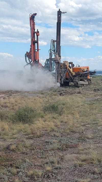 The image shows two large drilling rigs operating in an open, arid field under a cloudy sky. The rig on the left is orange, and the rig on the right is primarily orange and yellow. Both rigs are actively drilling, as indicated by the large cloud of dust or debris being generated at the base of the rig on the right. The terrain is covered in sparse, dry grass and rocky soil. In the distance, a body of water or a large expanse of land can be seen, possibly the city of Saran, Kazakhstan, which is known for its industrial activities, including mining. There are no people visible in the image. The overall scene depicts an industrial operation in a natural environment.