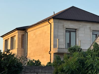 This image captures a close-up view of a large, modern house with a dark, tiled roof. The house is constructed with light-colored stone or concrete blocks, and features decorative detailing around the windows and eaves. Two sections of the house are visible: a two-story section with multiple windows on the left, and a larger section on the right with a balcony and a tall window. The building is illuminated by the warm, golden light of what appears to be either sunrise or sunset.
In the foreground, lush green trees and bushes partially obscure a low, grey stone wall. The foliage is dense, with a variety of leaf shapes and sizes, some of which are catching the golden light from the sky. A black gutter runs vertically down the side of the right section of the house, disappearing behind the trees. A small, dark chimney pipe protrudes from the peak of the roof.
The sky above is a clear, pale blue, indicating fair weather. The overall scene suggests a residential setting, likely in a suburban or semi-rural area given the presence of mature trees and the style of the house. The time of day is clearly either early morning or late evening due to the warm, directional sunlight casting long shadows and highlighting the architectural features of the building. There is no visible text in the image. The mood is peaceful and serene, enhanced by the soft light and the natural elements.