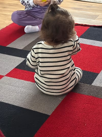 A child wearing a white and black striped sweater is sitting on a rug with red, grey, and black geometric patterns. The child has light brown hair. A second child wearing a white and black patterned long-sleeved shirt and purple pants is partially visible to the left, appearing to hold a yellow object. The scene is inside, likely on a wooden floor. Location information is not discernible from the image.