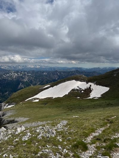 This is a high-angle, full shot of a mountainous landscape under a cloudy sky. The foreground features rough, grey rocks interspersed with green grass. A steep, grassy slope descends from the mid-ground to the lower right. In the mid-ground, a large patch of white snow lies on a green, undulating mountainside, partially melted and dark in places. Further into the background, layers of dark green, rugged mountains recede into the distance, some capped with snow. The farthest mountain range is hazy and appears to be under a lighter sky. The clouds are heavy and grey, with some patches of lighter sky visible. There are no people or specific man-made objects visible that would indicate a precise location within Jošanica, Montenegro, beyond it being a high-altitude mountainous area.