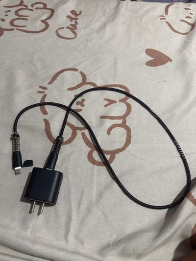 A black USB-C charging cable and adapter are laid out on a white, patterned fabric. The fabric features a repeating design of brown cartoon-like faces and hearts. The charging cable has a coiled section near the USB-C connector, which is a small, metallic connector with a black protective cap attached by a thin strap. The adapter is also black and has two metal prongs protruding from its base. The scene is lit from above, casting shadows on the fabric. The image is taken from a top-down perspective, with no specific location in Manila, Philippines identifiable.