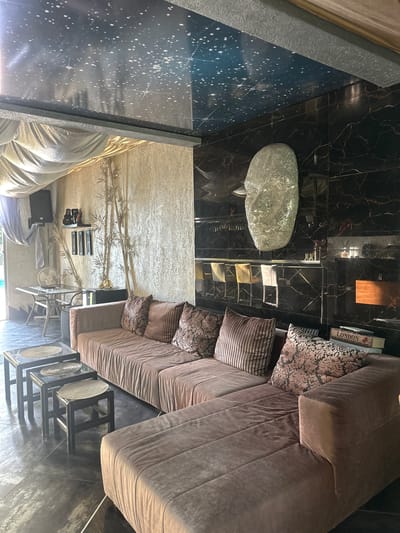 The image depicts a luxurious interior space, possibly a lounge or living room, in Brca, Montenegro. The room features a large, sectional sofa upholstered in a taupe velvet fabric, adorned with several decorative throw pillows in various patterns and textures. In front of the sofa, a set of three nested square glass-top tables with dark wood frames are arranged on a dark, reflective floor.
The backdrop of the room is striking. The ceiling is adorned with a dark, glossy finish patterned with white stars, resembling a night sky. One wall is covered in large, black marble tiles with prominent gold veining, creating a dramatic effect. Mounted on this wall is a large, textured sculpture of a human face, made of a reflective, possibly glitter-encrusted material. Adjacent to the marble wall is a textured, light-colored wall, decorated with several tall, stylized bamboo-like elements in a golden hue. On a shelf mounted on this wall are a few decorative items, including what appear to be speakers, a small golden figurine, and framed artwork. Further down this wall, a glass-top table with a metal frame is set up, with a fan and other objects on its surface, suggesting it might be a workspace or a functional area.
The lighting in the room is atmospheric, with a warm glow emanating from a wooden-mounted light fixture on the marble wall, and other ambient lighting sources not directly visible. The overall impression is one of opulence and modern, eclectic design.