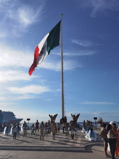 The image shows a group of people gathered in front of a flagpole in Cozumel, Mexico. The flagpole is located at the coordinates 20.511, -86.95. It is flying the Mexican flag, a tri-color of green, white, and red, with the national coat of arms in the center. The flag is waving in the wind, and the sky is blue with some clouds. The ground is paved, and there are some people standing around the flagpole. Some of the people are dressed in traditional Mexican clothing, while others are dressed in casual clothing. There are two large metal sculptures of birds on either side of the flagpole. In the distance, there is a cruise ship and a body of water. The scene is a festive gathering.