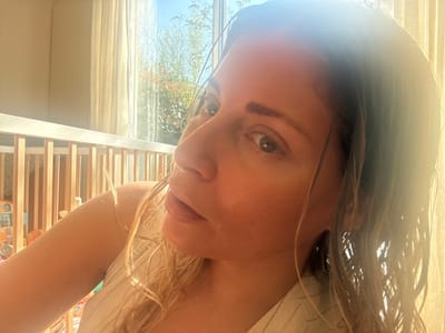An adult female is depicted in a close-up, eye-level portrait, her head tilted slightly as she looks directly towards the camera. Her light-colored hair, appearing wet, cascades over her shoulders. Strong natural light, originating from a window behind her, backlights the scene, illuminating her hair and skin with a warm glow and creating noticeable lens flare. The background features a large window covered partially by translucent, light-colored curtains. Beyond the window, green foliage and a clear blue sky are visible, indicating an outdoor garden setting during daytime. To the left of the frame, a segment of a light-colored wooden baby crib with vertical slats is present, with indistinct colorful objects inside. The scene is located within a room in Rueil-Malmaison, France.