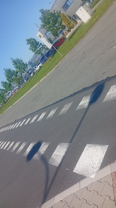 The image shows a street with white markings, likely a pedestrian crossing, in Karviná, Czechia. A shadow of a lamppost falls across the road. In the background, a modern building with windows and solar panels is visible, surrounded by trees and parked cars. The sky is clear and blue. The scene appears to be a sunny day in an urban or industrial area.