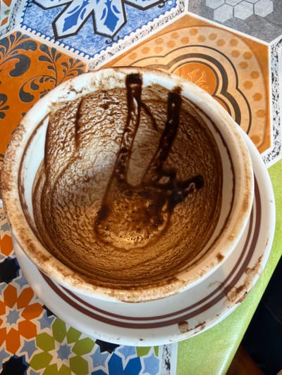 The image displays an overhead view of a white ceramic cup and saucer resting on a table surface in Eschau, France. The interior of the cup is coated with a substantial layer of dark brown, granular residue, consistent with the dregs of a consumed beverage such as coffee or hot chocolate. Streaks and patterns are visible within this residue, adhering to the inner walls and base of the cup. The exterior of the cup and a portion of the saucer also show smudges and drips of the same dark brown substance. The saucer is white ceramic with a thin brown band near its rim. The underlying surface is a brightly colored, patterned mat or tablecloth featuring a mosaic design composed of hexagonal and star-like shapes in various shades of blue, orange, green, and white. No individuals, direct actions, or interactions are visible in the scene.