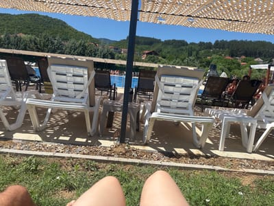 The image depicts an outdoor swimming pool area, likely a resort or public facility, with lounge chairs positioned under a sunshade structure. The pool is visible in the background, with a few people swimming or relaxing by its edge. Beyond the pool area, there are lush green hills covered in trees, and some houses are scattered on the slopes, suggesting a semi-rural or mountainous setting. The foreground shows the legs of at least two individuals, possibly sitting or lying on the grass, facing the pool and lounge chairs. The overall impression is a sunny day at a recreational spot, likely in Delchevo, North Macedonia, given the context.